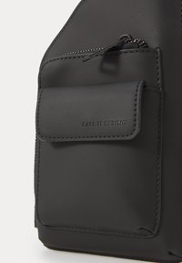 Zwarte rugzak met een matte afwerking, een ritszak en gestikte accenten, en een klepsluiting aan de voorkant met het merknaam in reliëf.