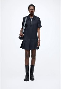 Vestito nero con colletto e maniche corte, con cerniera frontale e gonna arricciata. Abbinato a stivali neri al ginocchio.