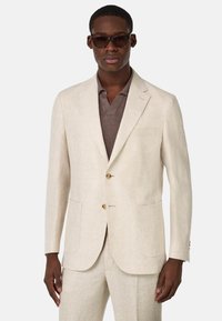 Blazer de lino beige claro con dos bolsillos frontales, diseño de un solo botón y botones marrones. Usado sobre una camiseta polo marrón oscuro.