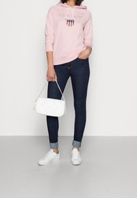 Jasnoróżowa bluza z kapturem ozdobiona logo i wzorem tarczy, w połączeniu z ciemnoniebieskimi dżinsami i białymi trampkami. Biała pikowana torebka trzymana w ręku.