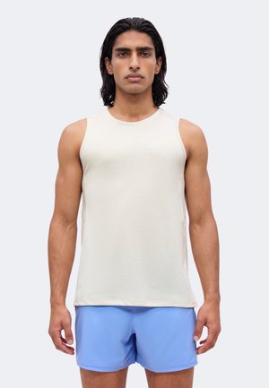 Homme aux cheveux foncés mi-longs, portant un haut beige sans manches et un short de sport bleu clair, debout devant un fond uni.