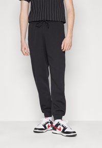 Černé tepláky s elastickým pasem, bočními kapsami a manžetami na kotnících; spárované s černým pruhovaným tričkem a černo-červenými teniskami.