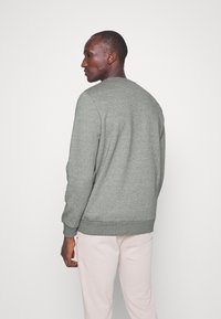 Felpa grigia con polsini e bordo a costine, caratterizzata da un colletto rotondo e una texture morbida. Indossata con pantaloni della tuta chiari. Vista posteriore mostrata.
