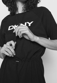 Svart klänning med en lös passform, elastiskt midjeband och en vit tryckt logotyp "DKNY" på framsidan. Tyget verkar mjukt med en avslappnad design.
