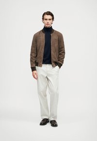 Young man standing wearing a brown suede jacket, black turtleneck, white trousers, and dark brown shoes on a white background.