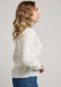 Blouse blanche brodée présentant des motifs floraux complexes, des manches bouffantes, des accents texturés et une finition transparente, associée à un jean bleu.