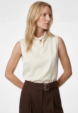 Mujer rubia que lleva una camiseta sin mangas de color crema con cuello abotonado y pantalones marrón oscuro de cintura alta con cinturón.