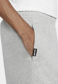 Pantalones de chándal grises con una textura suave, con bolsillos laterales y una etiqueta negra de "Just Do It" en el bolsillo. Diseño de corte casual.