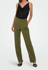 Žena v olivově zelených rovně střižených kalhotách, černém bezrukávovém topu s volným střihem a černých otevřených lodičkách na vysokém podpatku, stojící s jednou rukou v kapse.