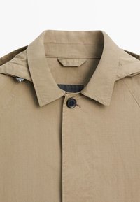 Beige trenchcoatkraag met één zwart knoopje dicht, met nette stiksels en een kleine stoffen lus onder de kraag.