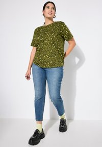 Camiseta verde con estampado de leopardo y mangas cortas, combinada con jeans azules. El modelo lleva zapatos negros de suela gruesa y calcetines verde claro.