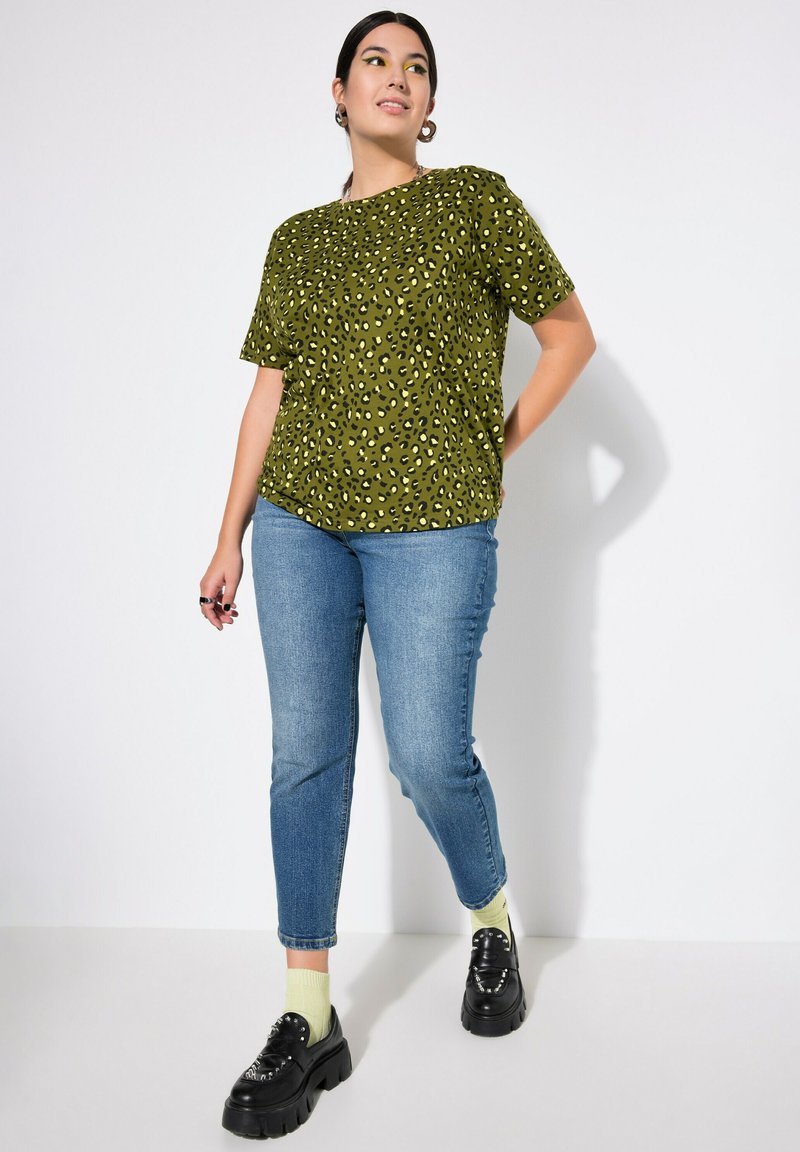 Camiseta verde con estampado de leopardo y mangas cortas, combinada con jeans azules. El modelo lleva zapatos negros de suela gruesa y calcetines verde claro.