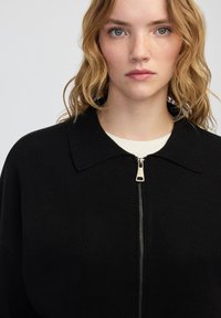 Blondýnka se vlnitými vlasy, oblečená v černé zipové bundě přes bílé tričko, hledí vpřed s neutrálním výrazem.