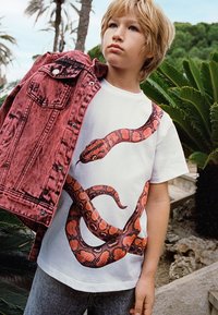 Rote Denimjacke, die über die Schulter eines Kindes hängt, mit einem weißen T-Shirt, das eine große, lebendige Schlangengrafik zeigt. Graue Jeans vervollständigen den Look.