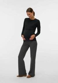 Majica s dugim rukavima za trudnice u crnoj boji, u kombinaciji s tamnosivim hlačama širokih nogavica. Jednostavan dizajn s mekim materijalom. Model drži stomak i stoji na ravnim cipelama.