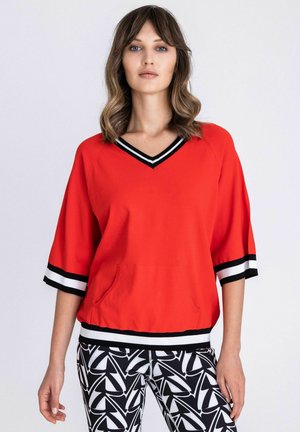 Vrouw draagt een rood V-hals topje met zwart-witte gestreepte manchetten en rand, gecombineerd met zwart-witte abstracte patroonbroek.