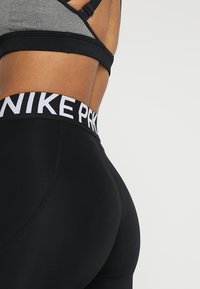 Svarta leggings med en bred vit midjeband som visar "NIKE PK"-logotypen. Slätt, elastiskt material. Grå sport-bh syns ovanf�ör leggings.
