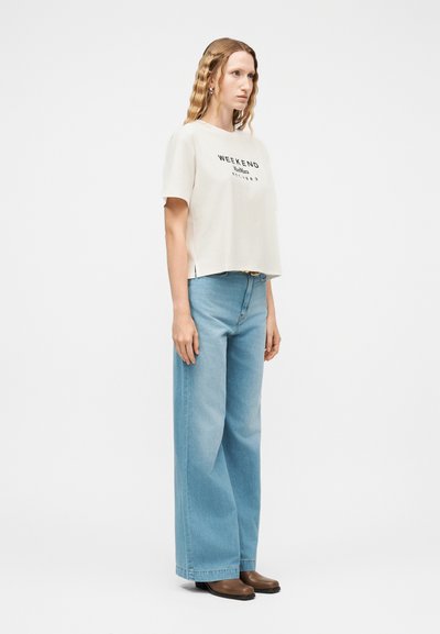 Γυναίκα με κυματιστά ξανθά μαλλιά φοράει λευκό μπλουζάκι με τη λέξη "WEEKEND", ανοιχτό μπλε τζιν παντελόνι με φαρδιά μπατζάκια και καφέ παπούτσια, στέκεται μπροστά σε απλό φόντο.