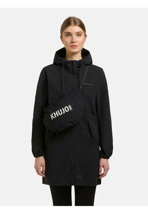 Kobieta w czarnym płaszczu z kapturem i czarnej torbie na ramię z białym napisem "KHUJO 2001", stojąca na jednolitym tle.