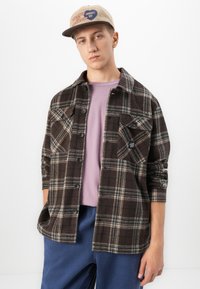 Jonge man met een beige pet, een bruine geruite getailleerde jas over een lila shirt en een blauwe broek, staand tegen een witte achtergrond.