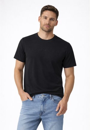 Uomo che indossa una t-shirt Richmond nera a maniche corte e jeans azzurri, in piedi con una mano in tasca contro uno sfondo bianco.