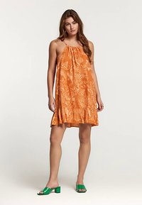 Oranges Kleid mit Blumenmuster, Rüschen-Saum und verstellbarem Neckholder-Ausschnitt. Komplettiert mit grünen Sandalen mit Blockabsatz.