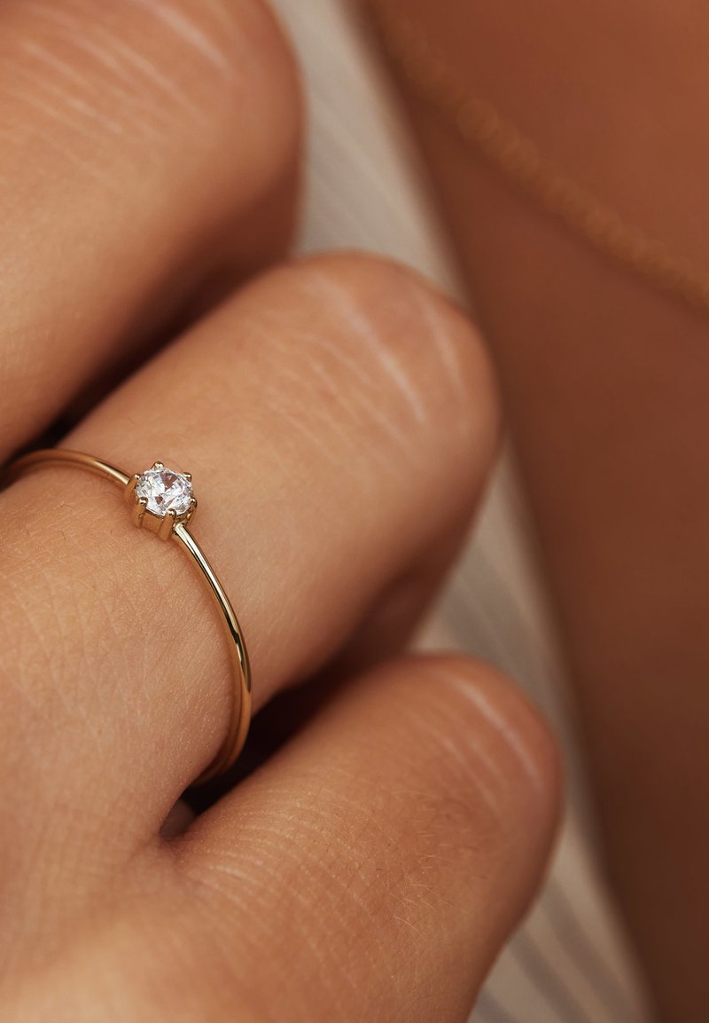 Goldring mit einem einzigen runden, klaren Edelstein, der über ein glattes, schlankes Band und eine filigrane Fassung verfügt. Der Finger zeigt einen hellen Hautton.