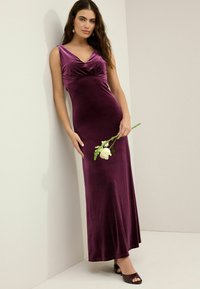 Robe en velours, longue jusqu'au sol, de couleur violet profond, avec un décolleté en V, un corsage froncé et une finition lisse. Le mannequin tient une rose blanche dans une main.