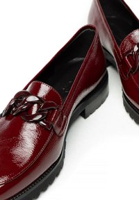 Glanzende donkerrode loafers met stevige zwarte zolen en een grote kettingdetail over de bovenkant op een witte achtergrond.