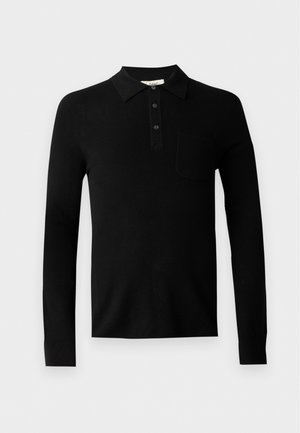 Homme debout portant un polo noir à manches longues, un pantalon marron ample et des chaussures noires sur un fond uni gris clair.