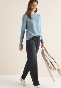 Hellblauer, gerippter Pullover, dunkelgraue weit geschnittene Jeans und weiße Schuhe mit roten Akzenten. Beigefarbene Jacke in der Hand. Einfaches, lässiges Outfit.