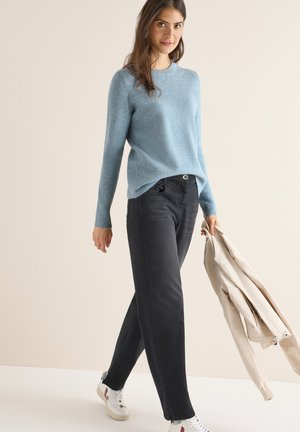 Hellblauer, gerippter Pullover, dunkelgraue weit geschnittene Jeans und weiße Schuhe mit roten Akzenten. Beigefarbene Jacke in der Hand. Einfaches, lässiges Outfit.