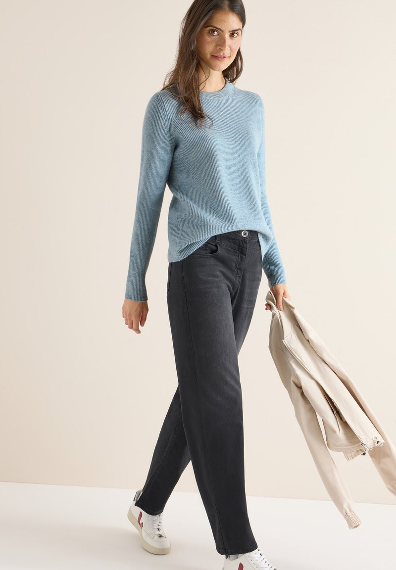 Hellblauer, gerippter Pullover, dunkelgraue weit geschnittene Jeans und weiße Schuhe mit roten Akzenten. Beigefarbene Jacke in der Hand. Einfaches, lässiges Outfit.