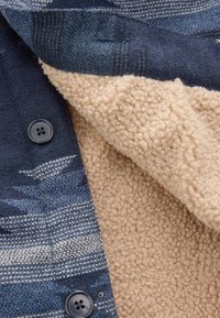 Blue and beige jacket featuring a textured fleece lining, striped wool exterior, and black button detail. Soft and warm material contrast.