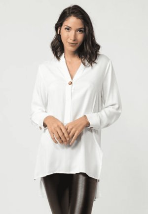 Blusa blanca de manga larga con botonadura frontal, con cuello y espalda ligeramente más larga, combinada con leggings negros. Textura suave.