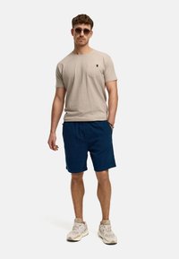 T-shirt beige à manches courtes avec poche, associé à un short bleu marine. Le mannequin porte des baskets grises et des lunettes de soleil, arborant une coupe décontractée et relaxée.