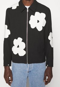 Chaqueta negra con grandes diseños florales blancos, cierre de cremallera frontal, cuello con puntas y bolsillos laterales. La tela parece ligera con una textura suave.