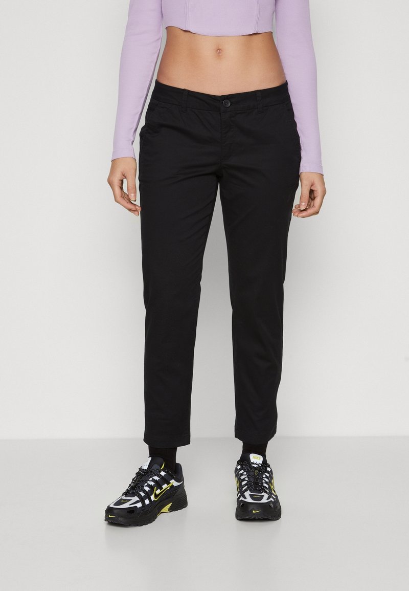 Femme portant un pantalon noir droit, des baskets noires et jaunes, et un top court lavande à manches longues, debout devant un fond blanc.