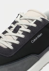 Calvin Klein sneaker with light gray laces, black and gray suede panels, beige tongue, and white rubber sole.