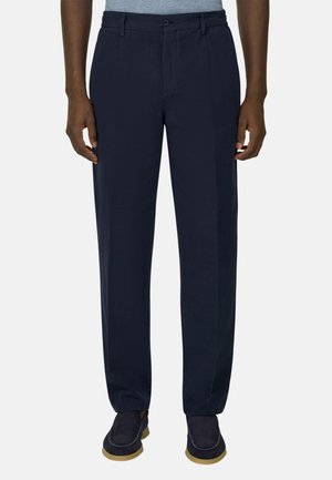 Homme portant un pantalon ajusté bleu marine, une chemise grise rentrée, et des chaussures en daim bleu foncé avec des semelles marron clair, debout droit sur fond blanc.