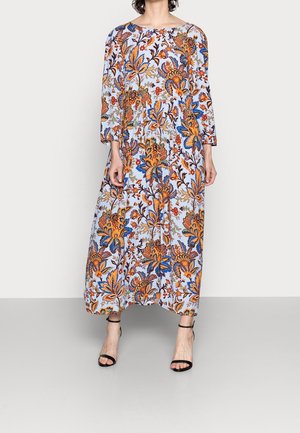 Mujer con un vestido largo y suelto de flores con patrones en naranja, azul y marrón, combinado con zapatos de tacón negros con tiras en el tobillo y puntera abierta.