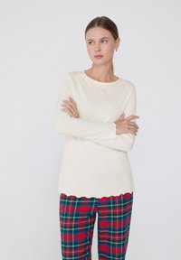 Blusa de manga larga color crema con un escote redondeado y dobladillo en punta, combinada con pantalones de cuadros rojos y verdes que presentan un patrón de cuadros clásico.