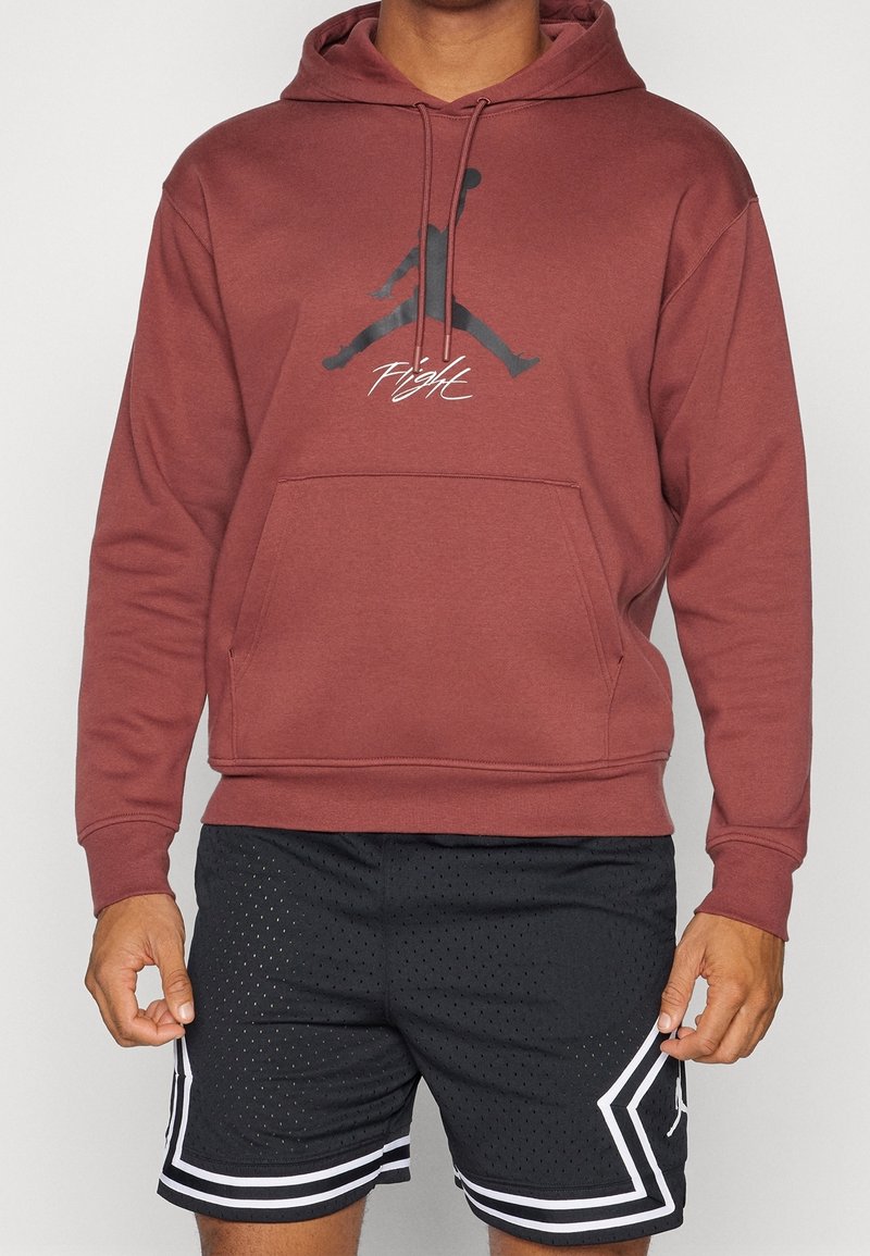 Personne portant un hoodie bordeaux avec un logo Jumpman noir et un texte blanc "Flight", associé à un short de sport noir perforé avec des rayures blanches.