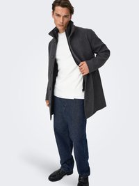 Grauer Wollmantel mit hohem Kragen, Knopfverschluss und kontrastierendem Innenfutter, getragen über einem weißen, strukturierten Pullover und dunkelblauen Jeans.