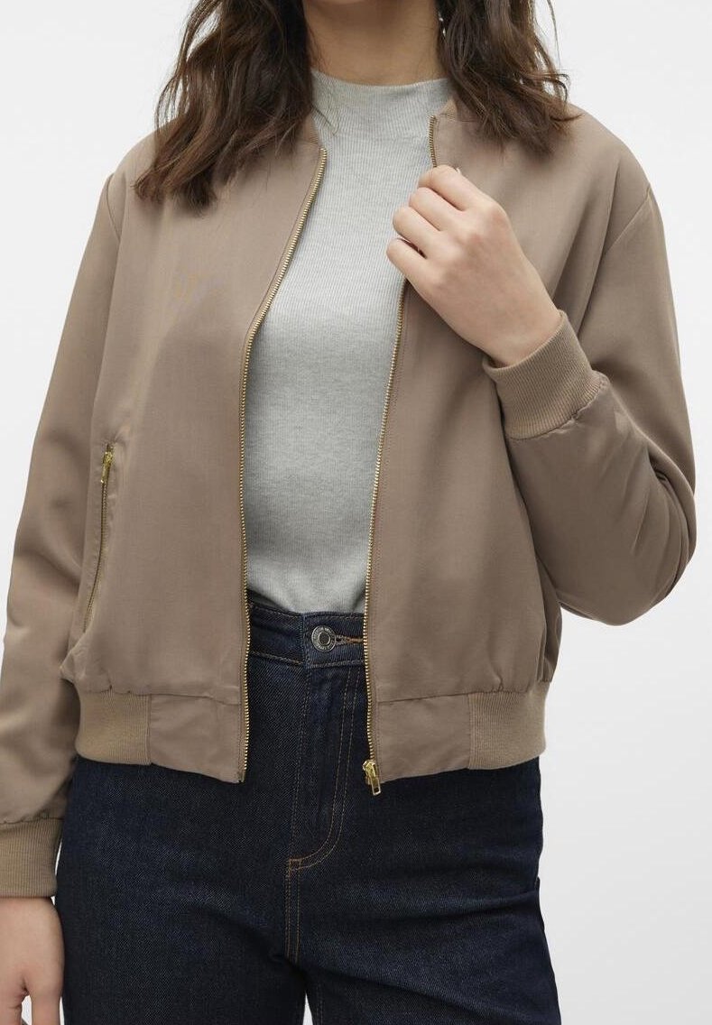 Femme portant une veste bomber beige zippée sur un haut gris clair et un jean taille haute bleu foncé, main près de la fermeture éclair de la veste.