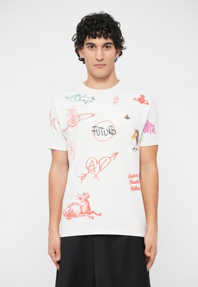 Jovem com cabelo encaracolado, vestindo uma t-shirt branca com desenhos e texto em vermelho, rosa e verde, combinada com calças pretas, de pé contra um fundo branco.