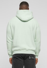 Felpa verde menta con tasca a marsupio, cappuccio con cordino e polsini a costine; indossata sopra jeans scuri con un logo ricamato bianco sulla tasca posteriore.