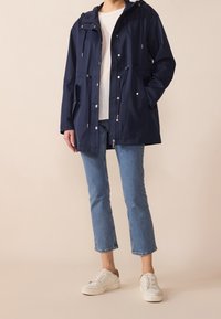 Marinblå vattentät jacka med huva, främre dragkedja, dragsko, sidofickor och silverdetaljer. Kombination med ljusblå jeans och vita sneakers.