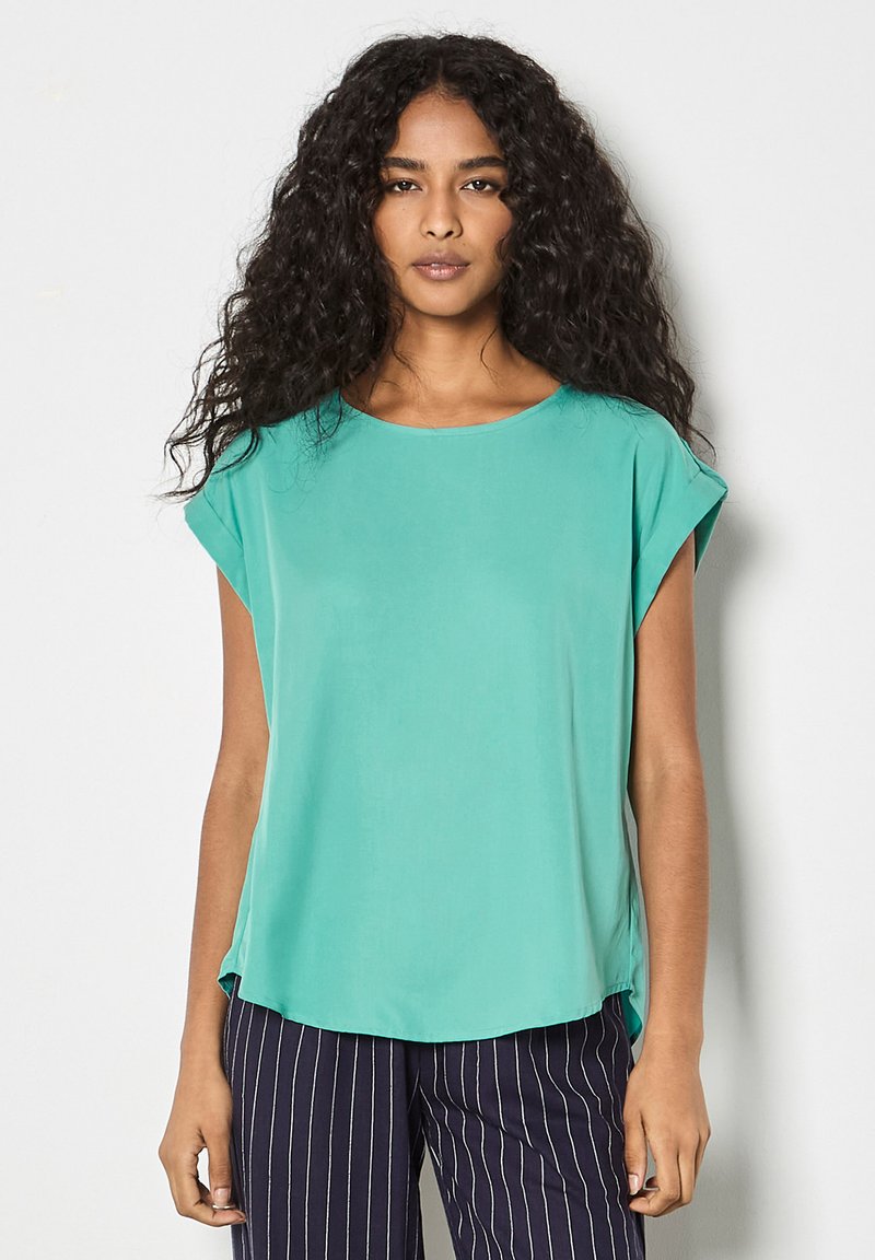 Femme aux longs cheveux bouclés portant un chemisier turquoise à manches courtes amples et un pantalon bleu marine à rayures verticales blanches sur un fond uni.