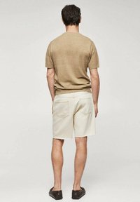 Pull court en tricot beige, short en crème avec bord effiloché, et chaussures foncées. Vu de dos contre un fond gris clair.